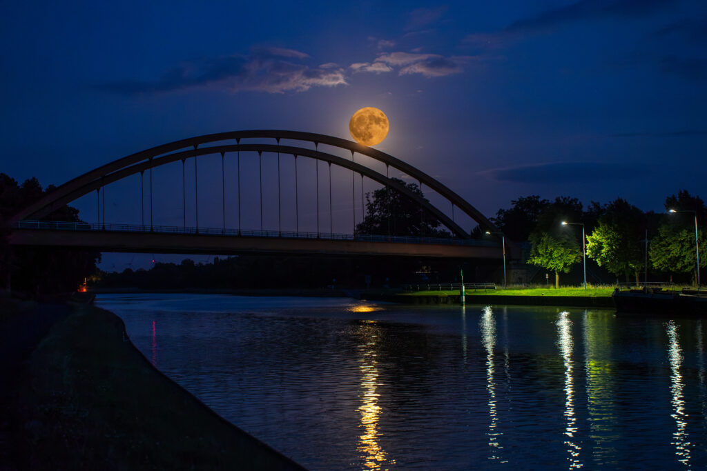 Der große Vollmond leuchtet in warmem Gold, eingerahmt von einer markanten Bogenbrücke über einem ruhigen Fluss. Die Spiegelungen der Lichter tanzen im Wasser, während der Nachthimmel in tiefem Blau leuchtet. Die Brücke teilt das Bild elegant, während Bäume und Laternen eine sanfte Szenerie bilden.